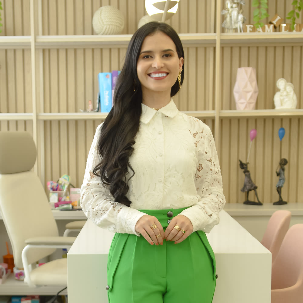 Dra. Thamires Mattos em pé no consultório, apoiada na mesa, com estantes e poltronas rosadas ao fundo