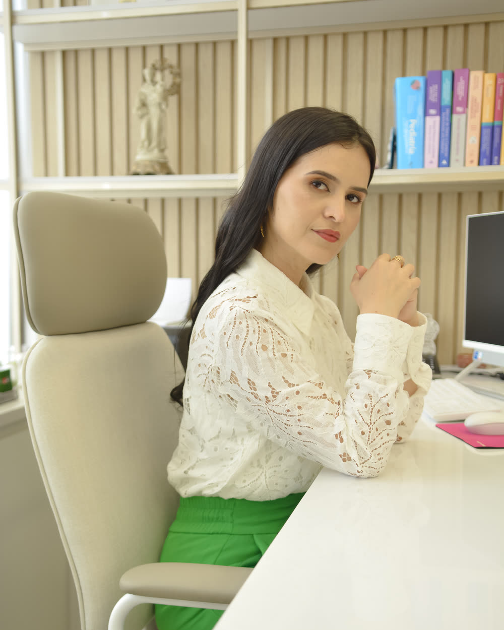 Dra. Thamires Mattos sentada à mesa do consultório, de blusa de renda branca, olhando para a câmera